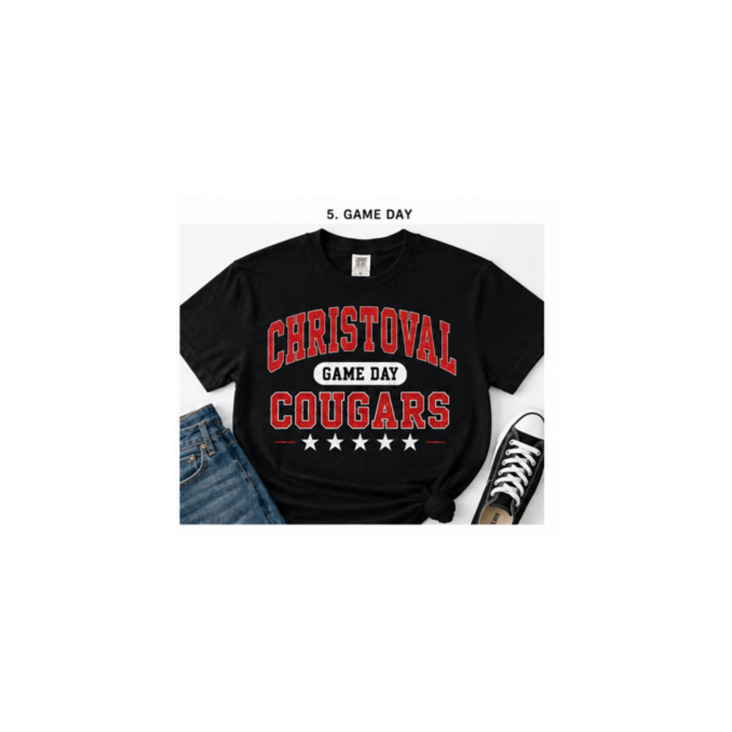 A black short-sleeve graphic T-shirt laid flat with blue jeans and a black sneaker as styling props. The shirt has large red collegiate-style text reading 'CHRISTOVAL GAME DAY COUGARS' with white stars and a white banner reading 'GAME DAY'.