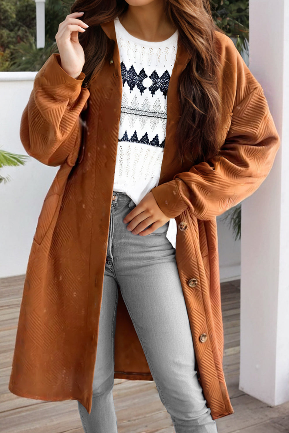 Person wearing a brown coat over a white sweater with black patterns, standing on a wooden deck.
