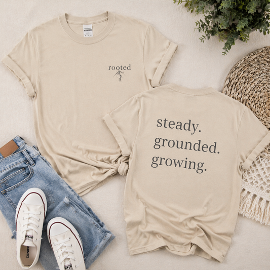 Beige t-shirt with motivational text rooted on front steady grounded growing on back , paired with jeans and sneakers on a light surface.
